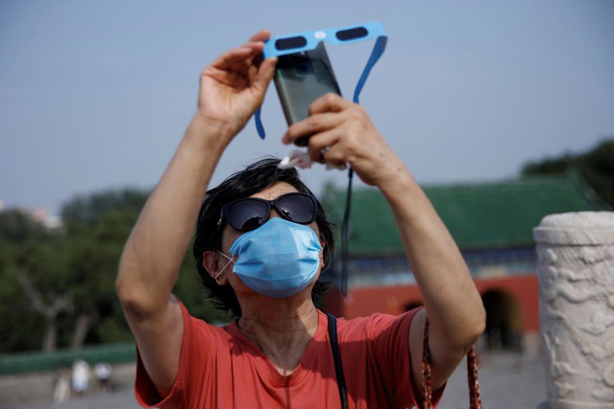 Eclipse solaire : une femme portant un masque en raison de la pandémie de coronavirus ou tenant des lunettes de protection devant son téléphone alors qu'elle capture l'éclipse partielle de soleil dans le parc du Temple du Ciel à Pékin.