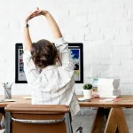 Une femme s'étire sur sa chaise de bureau devant son ordinateur, illustrant l'importance d'une bonne posture pour le travail prolongé.