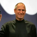 Steve Jobs tenant le premier iPhone lors de sa présentation officielle, vêtu de son col roulé noir iconique.