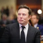 Portrait d'Elon Musk lors d'une conférence de presse, regard concentré vers la droite, en costume-cravate sombre.