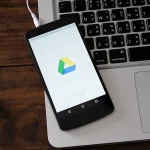 Logo de l'application Google Drive affiché sur l'écran d'un smartphone Android, posé sur le clavier d'un ordinateur portable. Illustration de l'accessibilité et de la synchronisation des données via le service de stockage cloud.