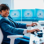 Un adolescent en costume bleu tape sur un clavier d’ordinateur, entouré de kits STEM et de composants électroniques, dans un bureau lumineux.