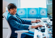 Un adolescent en costume bleu tape sur un clavier d’ordinateur, entouré de kits STEM et de composants électroniques, dans un bureau lumineux.