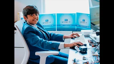 Un adolescent en costume bleu tape sur un clavier d’ordinateur, entouré de kits STEM et de composants électroniques, dans un bureau lumineux.