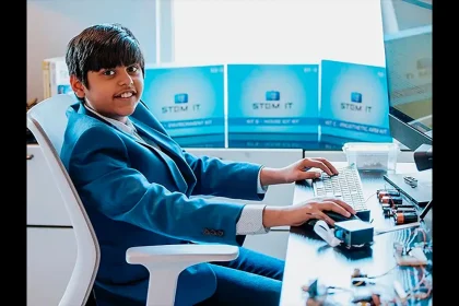 Un adolescent en costume bleu tape sur un clavier d’ordinateur, entouré de kits STEM et de composants électroniques, dans un bureau lumineux.