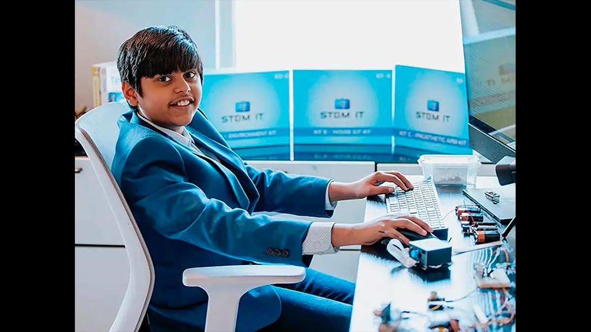 Un adolescent en costume bleu tape sur un clavier d’ordinateur, entouré de kits STEM et de composants électroniques, dans un bureau lumineux.
