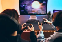Deux joueurs portant des casques audio jouent à un jeu vidéo sur un écran PC à l’aide de manettes, devant un bureau lumineux.