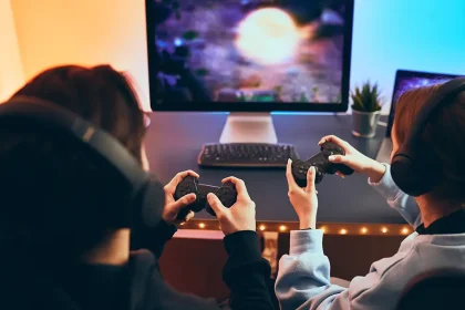 Deux joueurs portant des casques audio jouent à un jeu vidéo sur un écran PC à l’aide de manettes, devant un bureau lumineux.