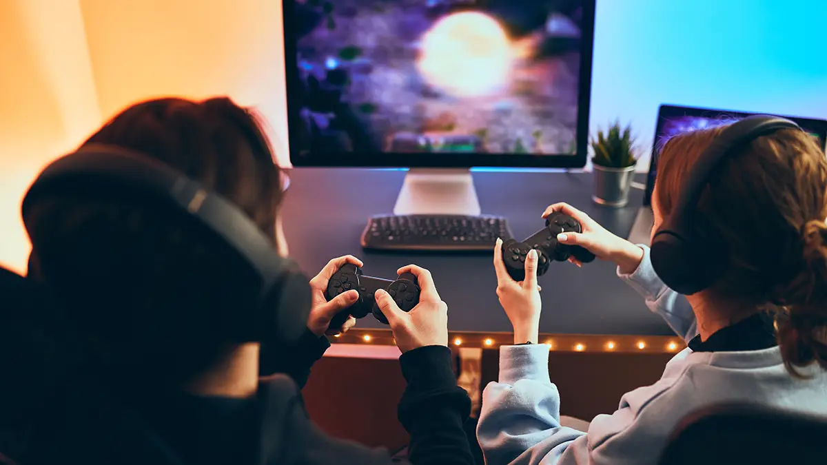 Deux joueurs portant des casques audio jouent à un jeu vidéo sur un écran PC à l’aide de manettes, devant un bureau lumineux.