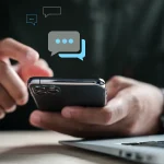 Main tenant un smartphone, écran affichant des bulles de messages SMS, devant un ordinateur portable