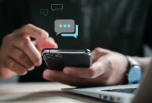 Main tenant un smartphone, écran affichant des bulles de messages SMS, devant un ordinateur portable