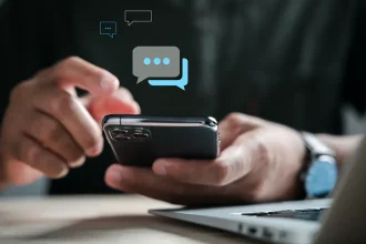 Main tenant un smartphone, écran affichant des bulles de messages SMS, devant un ordinateur portable