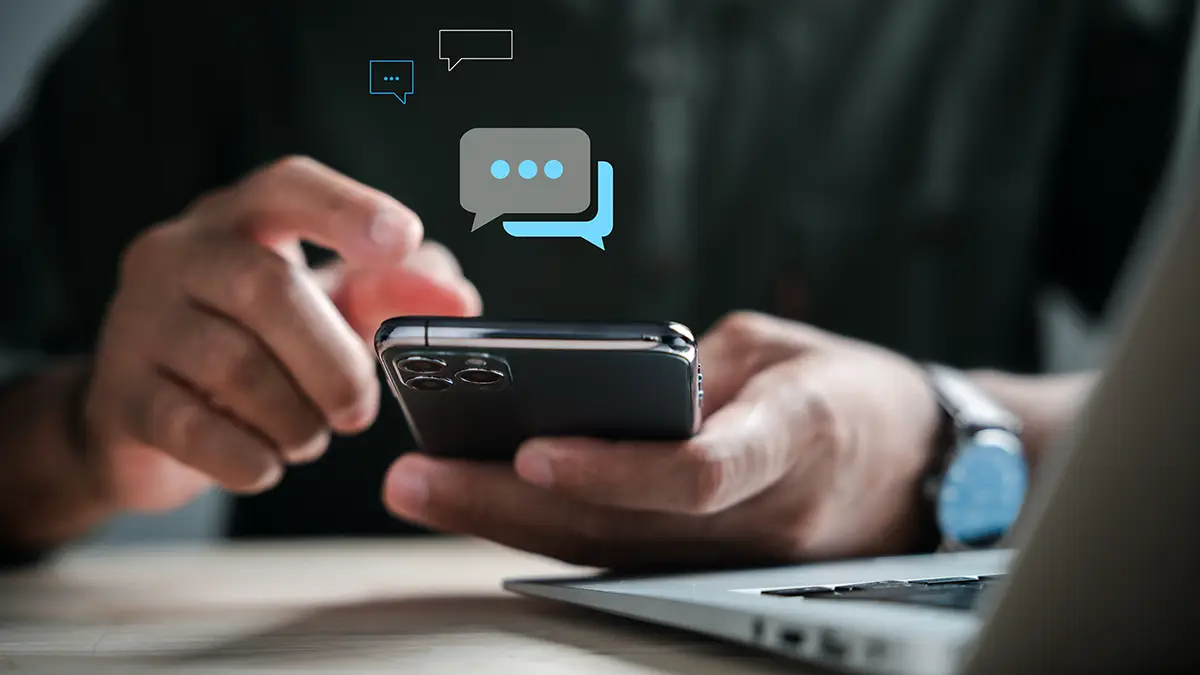 Main tenant un smartphone, écran affichant des bulles de messages SMS, devant un ordinateur portable