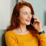 Jeune femme souriante assise sur un canapé, en train de téléphoner dans un intérieur lumineux