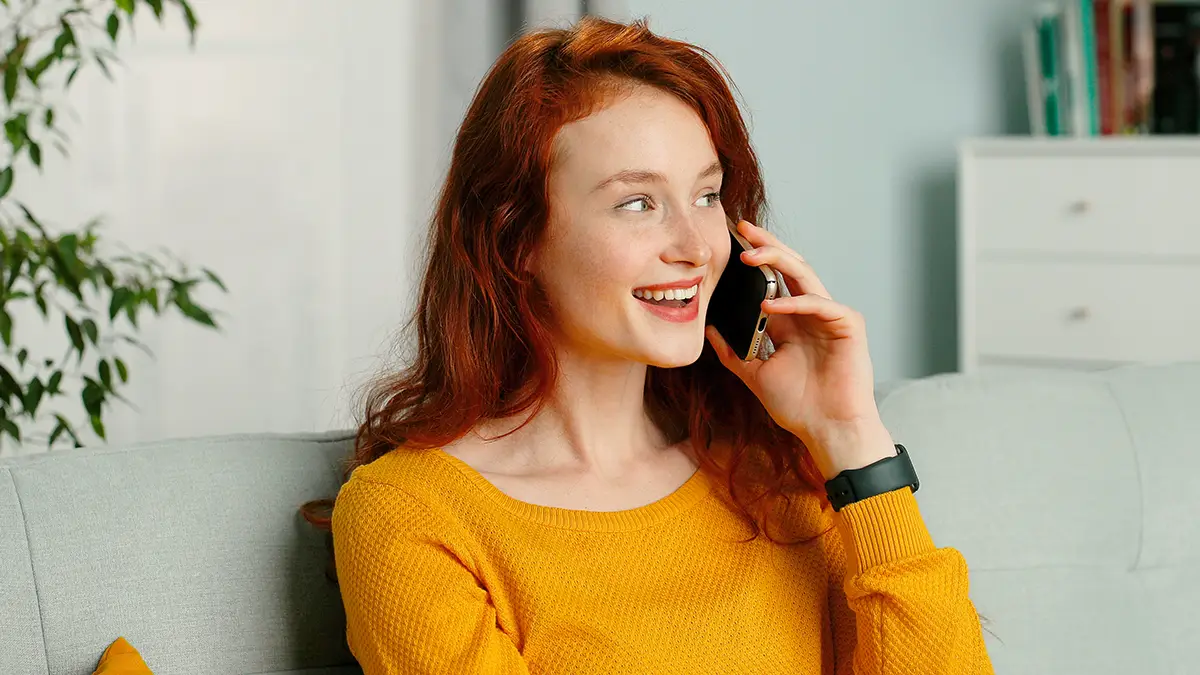 Jeune femme souriante assise sur un canapé, en train de téléphoner dans un intérieur lumineux