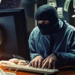 Un individu cagoulé devant un écran d’ordinateur, entouré de billets de banque, symbolisant une cyberattaque de type jackpotting sur distributeur automatique.