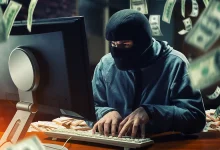 Un individu cagoulé devant un écran d’ordinateur, entouré de billets de banque, symbolisant une cyberattaque de type jackpotting sur distributeur automatique.