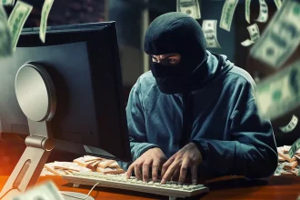 Un individu cagoulé devant un écran d’ordinateur, entouré de billets de banque, symbolisant une cyberattaque de type jackpotting sur distributeur automatique.