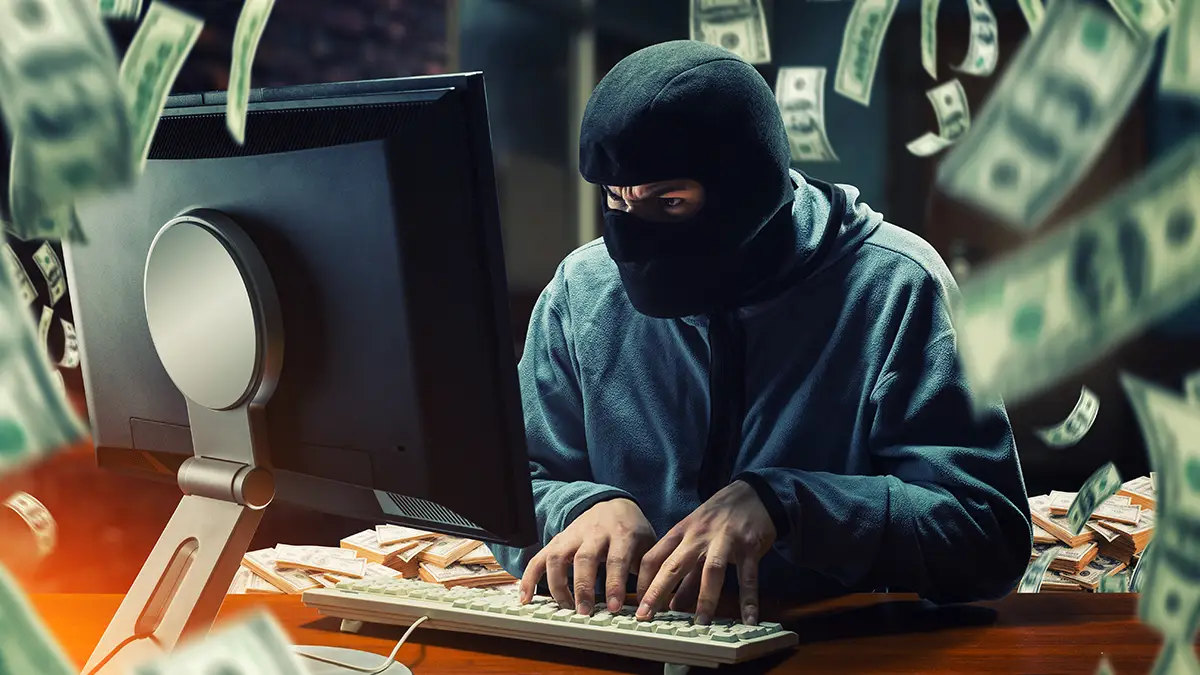 Un individu cagoulé devant un écran d’ordinateur, entouré de billets de banque, symbolisant une cyberattaque de type jackpotting sur distributeur automatique.